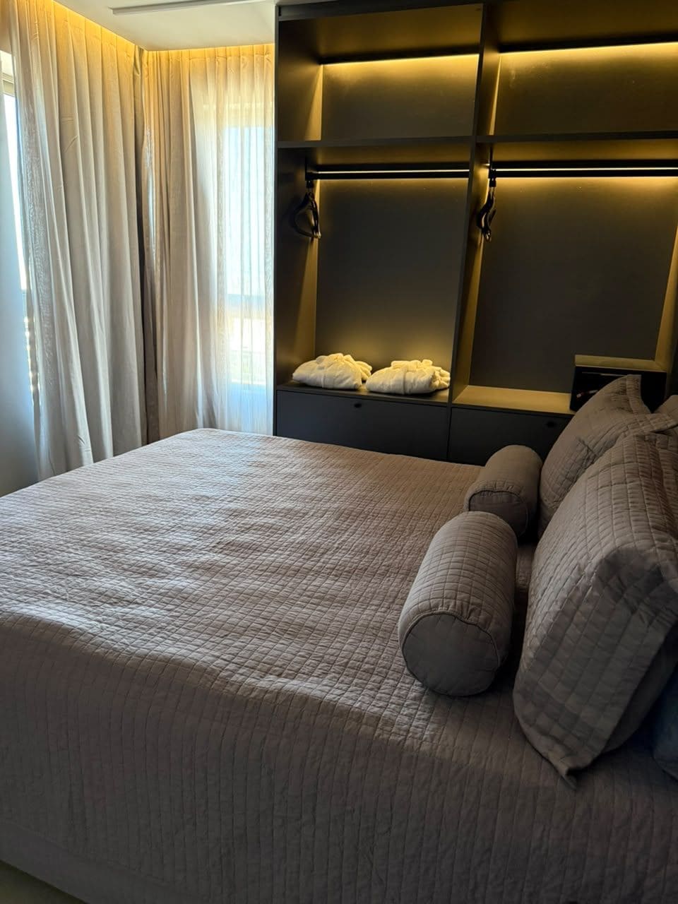 Una cama bien hecha con cubrecama acolchado, cabecera con iluminación integrada y mesita de noche con toallas.