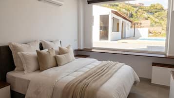 A bedroom with a large bed, a window with a view of a building and a pool, and a white wall.