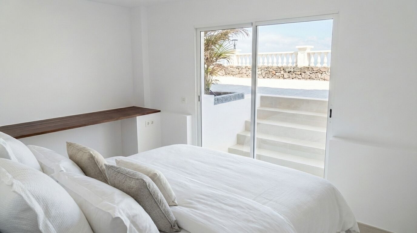 Une chambre à coucher avec un lit, des oreillers, une étagère en bois et une porte-fenêtre coulissante donnant sur un balcon extérieur agrémenté d’un mur en pierre et de palmiers.