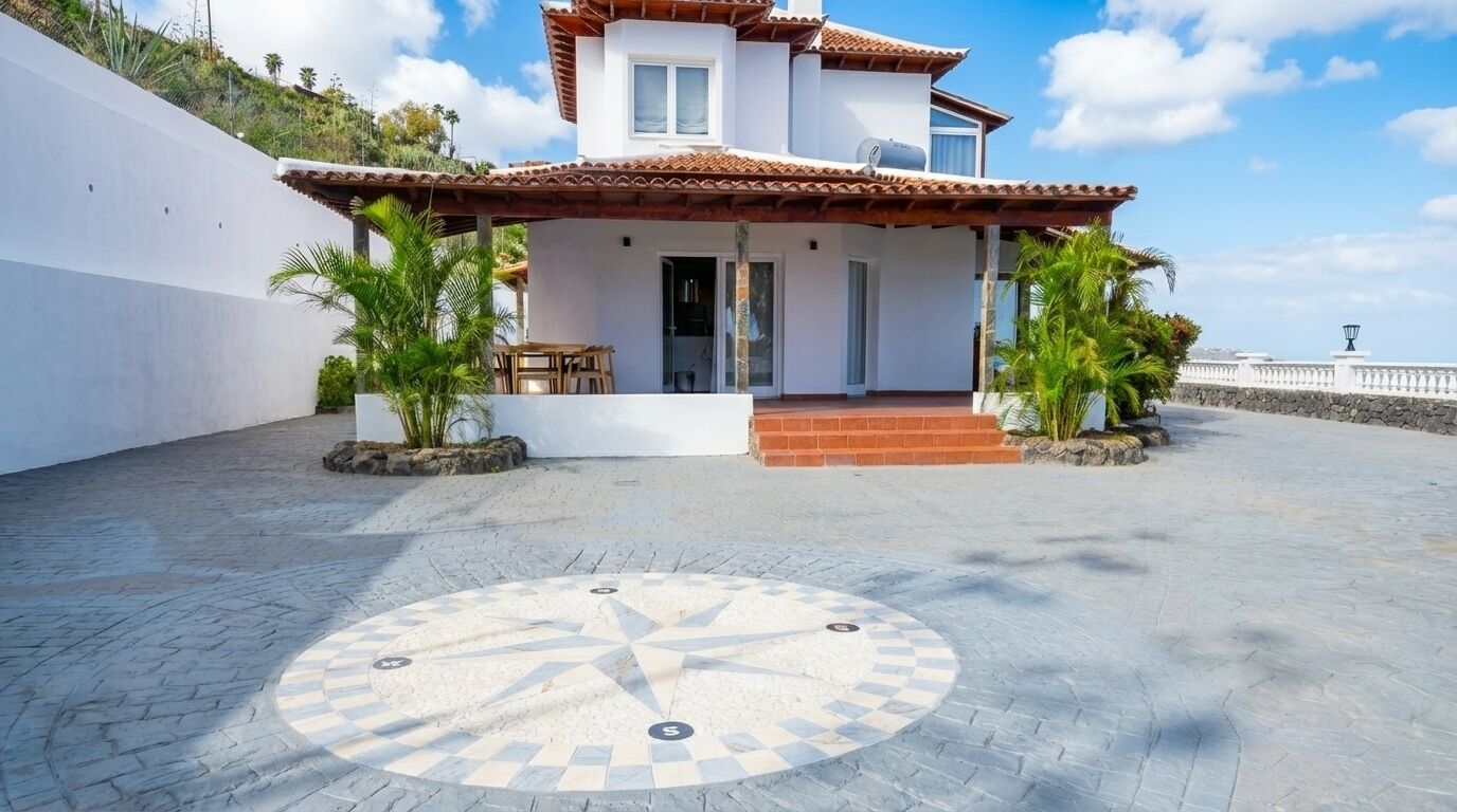 A two-story house with a tiled roof, a paved courtyard with a compass design, and a white wall on one side.