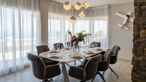 A dining area with a round table set for six, surrounded by chairs, with a view of the outdoors through large windows. - Exclusive Mountain Villa (Barranco Hondo)