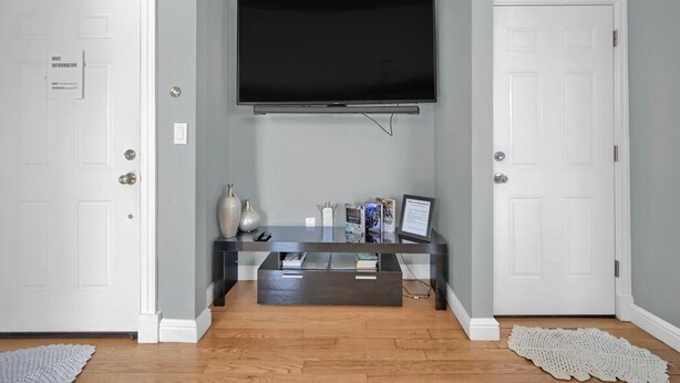 Een kamer met een wandgemonteerde flatscreen-tv, een donker houten salontafel met tijdschriften en een laptop, en een witte deur met een bordje.
