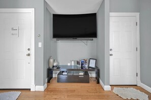 A room with a flat-screen TV mounted on the wall, a dark wooden coffee table with magazines and a laptop, and a white door with a sign. - Enjoyable 3-bedroom apartment with AC in vibrant Somerville (Somerville)