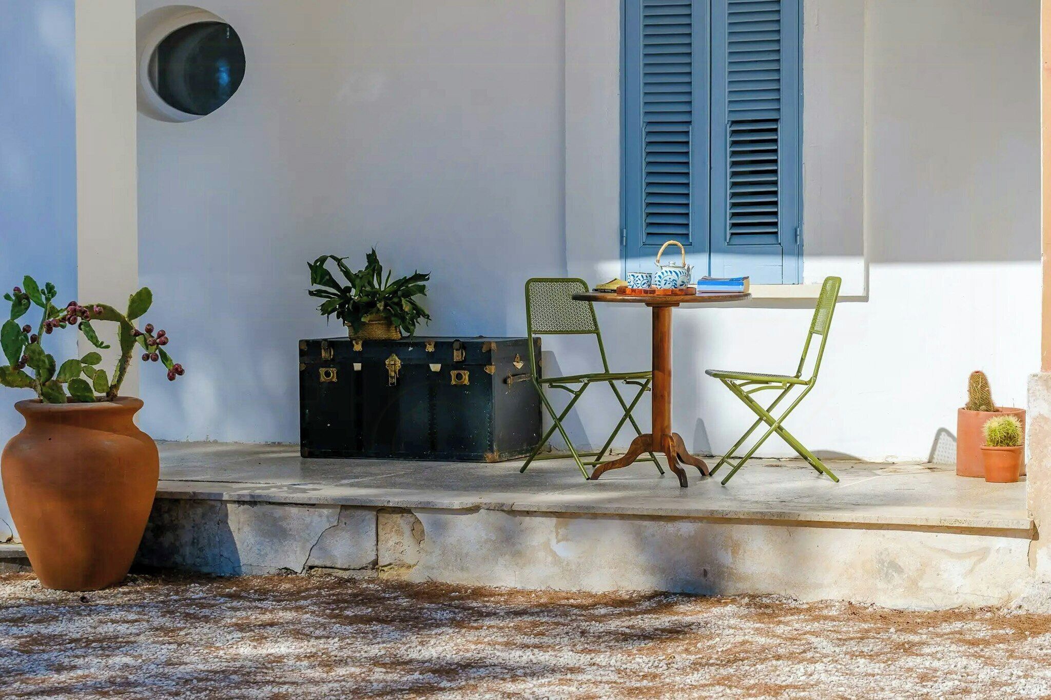 Una terrazza con tavolo, sedie, una pianta in vaso, una valigia e una finestra con persiane.