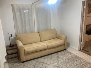 Ein beiges Sofa in einem Raum mit einem gemusterten Teppich, einem kleinen Tisch mit Lampe und einer teilweise sichtbaren Tür.