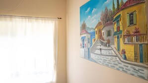 Ein Zimmer mit einem großen Fenster, das mit einem weißen Vorhang bedeckt ist, einem Wandbild einer farbenfrohen Straßenszene und einer weißen Decke mit einem sichtbaren Stromanschluss.