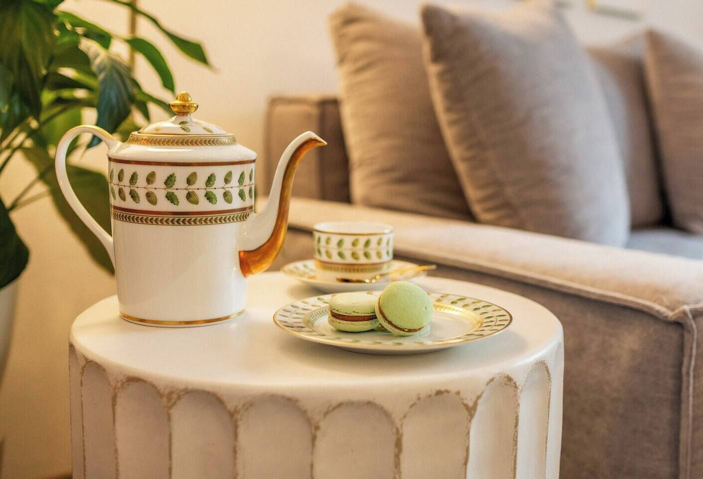Valkoinen pöytä, jonka päällä on koristeellinen teekuppisetti, johon kuuluu teekannu ja kuppeja, sekä kaksi vihreää macaron-leivosta lautasella.