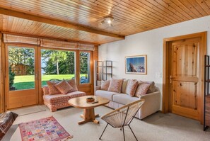 A cozy living room with a wooden ceiling, two sofas with patterned cushions, a round wooden coffee table, a wicker chair, and a door leading outside.