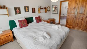 Ein ordentlich bezogenes Bett mit weißen Leinen und Karokissen, ein Nachttisch aus Holz mit einer Lampe, ein kleiner Holztisch und ein Kleiderschrank mit Spiegel.