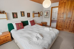 Ein ordentlich bezogenes Bett mit weißen Leinen und Karokissen, ein Nachttisch aus Holz mit einer Lampe, ein kleiner Holztisch und ein Kleiderschrank mit Spiegel.