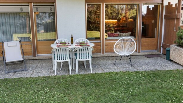 Eine Terrasse mit Tisch für vier Personen, umgeben von weißen Stühlen, und ein einzelner Stuhl außerhalb der Terrasse.