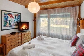 A bedroom with a bed, a wooden dresser, a television with a fire, and a window with curtains.