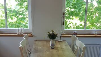 Et spiseområde med et træbord, hvide stole, en hængende pendel og et vindue med udsigt til frodig vegetation.