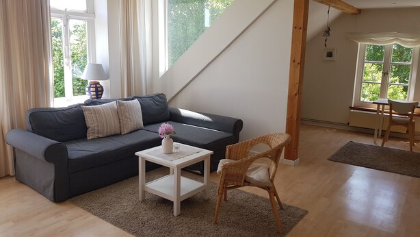 En hyggelig stue med en mørkegrå sofa, et lille hvidt bord med en vase, en fletstol og udsigt til det fri gennem store vinduer.