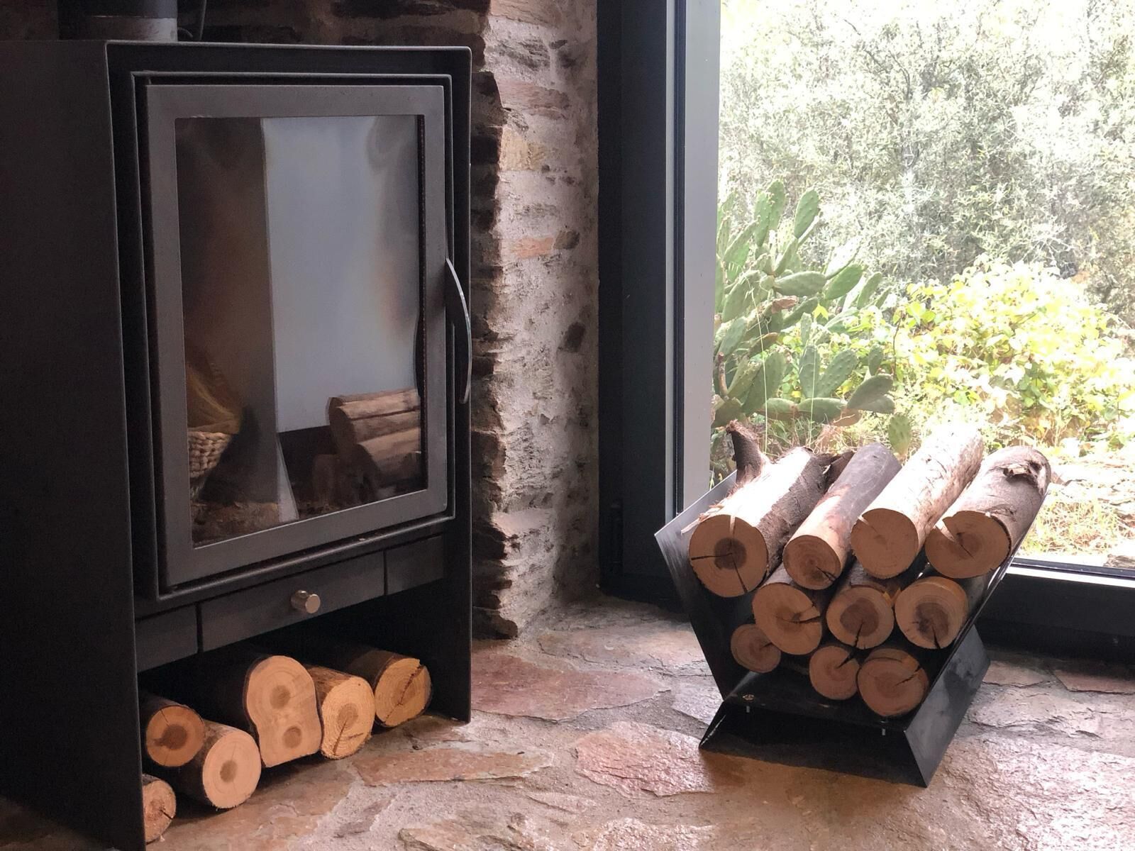 A fireplace with logs stored below and to the side, set against a stone wall with a window overlooking greenery.