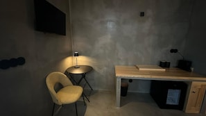 A small, dimly lit room with a single chair, a desk with a lamp, and a television mounted on the wall.