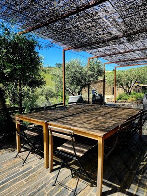 An outdoor dining area with a wooden table and chairs, a pergola overhead, and a view of a garden with trees and a stone wall in the background.