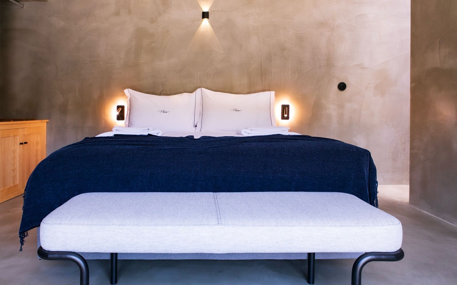 A bed with a dark blue cover, white pillows, and a white bench in front.