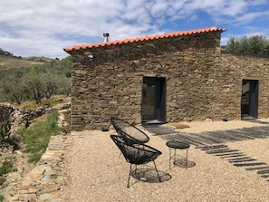 A stone building with a red-tiled roof, two doors, and a patio area with a chair and table.