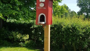 Una casetta per uccelli in legno con tetto rosso e un piccolo ingresso, fissata a un palo di legno decorato con nastri colorati.