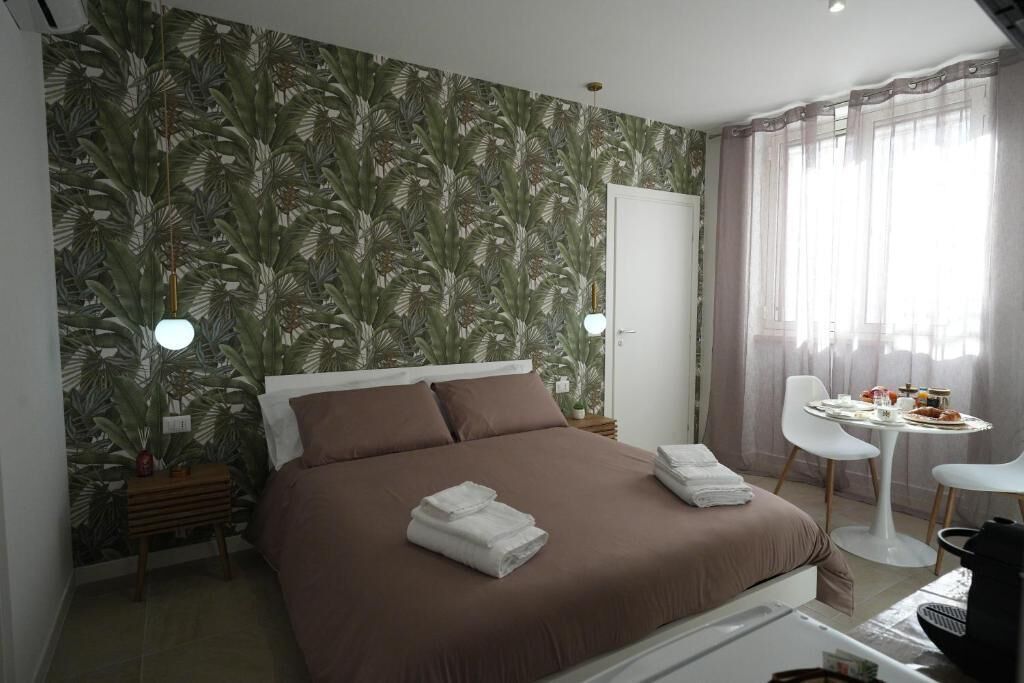 Une chambre à coucher comprenant un lit, une table de chevet, une chaise, une petite table avec quelques objets, et une fenêtre avec des rideaux.