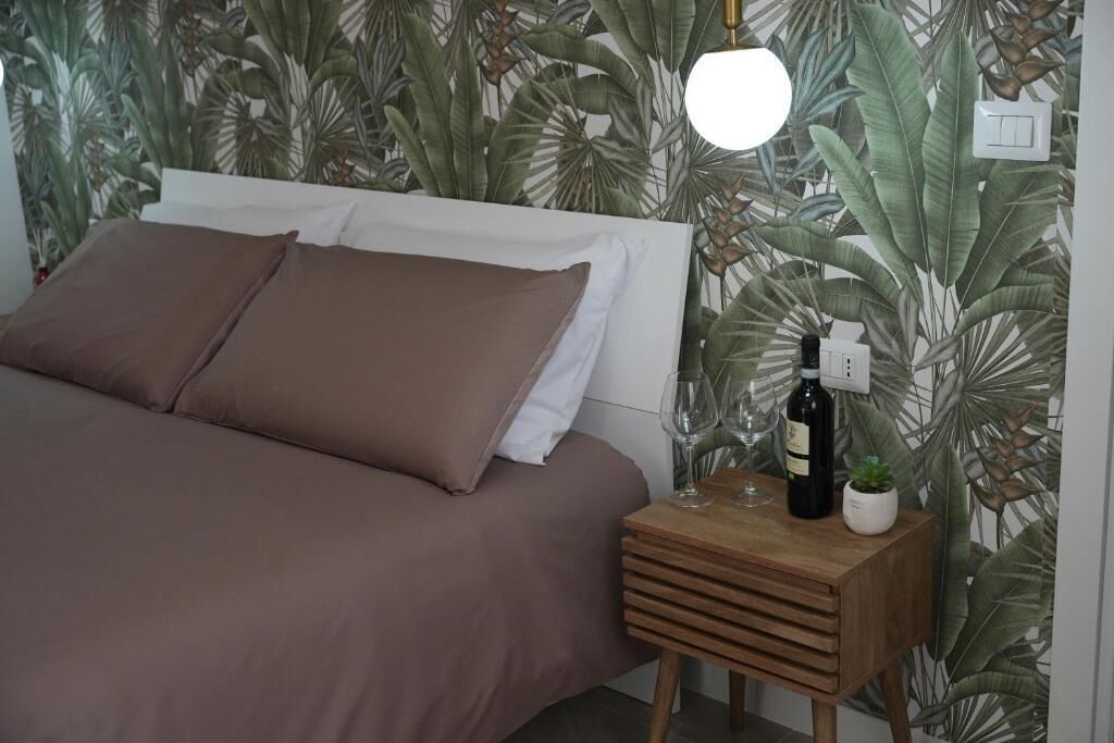 Un dormitorio con una cama, una mesita de noche con una botella y vasos, y una pared con un estampado de hojas tropicales.