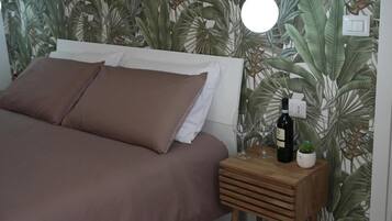 Un dormitorio con una cama, una mesita de noche con una botella y vasos, y una pared con un estampado de hojas tropicales.
