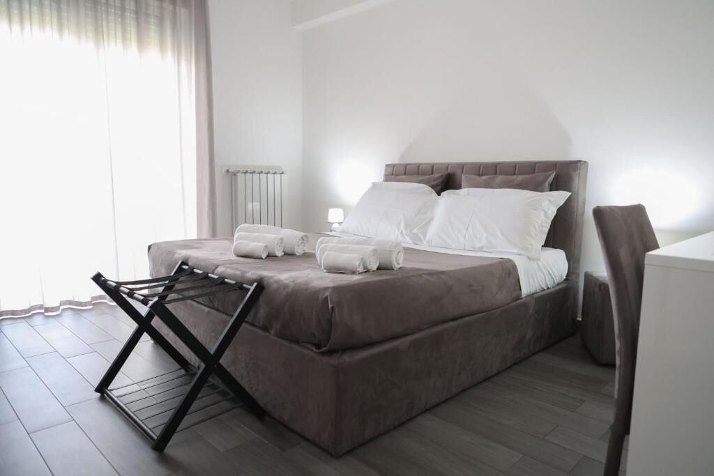 Un dormitorio con cama, almohadas, un radiador, una silla y una ventana con cortinas.