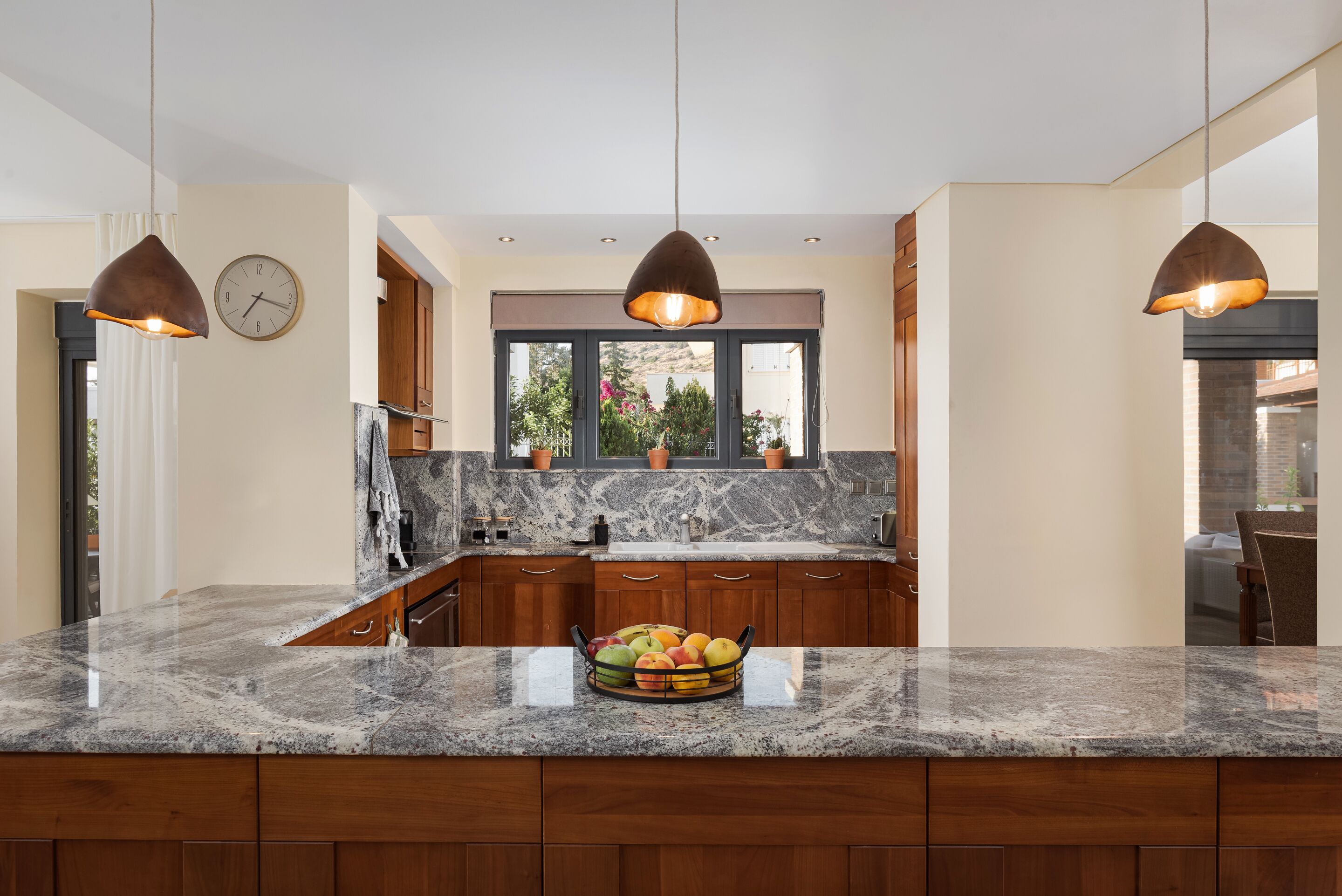 A kitchen with granite countertops, wooden cabinets, pendant lighting, a window with a view, and a fruit bowl on the counter. - Villa Liberta Athenian Riviera (Saronikos)