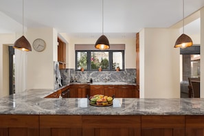Una cocina con encimeras de granito, gabinetes de madera, iluminación colgante, un ventanal con vistas y un frutero sobre la encimera.