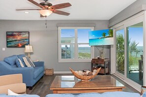 Ein Wohnzimmer mit einem blauen Sofa, einem Glastisch, einem Deckenventilator, einem Fernseher und einem großen Fenster mit Blick auf den Strand.
