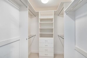 Ein begehbarer Kleiderschrank mit mehreren Regalen und Schubladen, einer Deckenleuchte und einer Tür links.