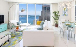 A modern living room with a sofa, armchair, dining table, and a large window with a view of the sea and buildings. - Icon Brickell 2BR · Panoramic Bay Views, Sleeps 6 (Miami)