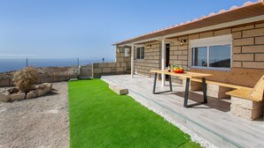 Eine moderne Terrassengrundstueck mit einem Holztisch und Sitzgelegenheiten, ein kleiner Gartenbereich mit Kunstgrasmattierung und Blick aufs Meer.