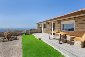 En moderne udendørs terrasse med et træbord og siddepladser, et lille haveområde med kunstgræs og udsigt til havet.