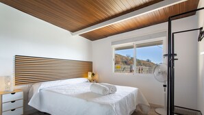 Ein Schlafzimmer mit einem Lattenrost, einem Bett mit weißer Bettwäsche, einem Nachttisch mit Lampe und einem Fenster mit Blick auf eine Hügellandschaft.