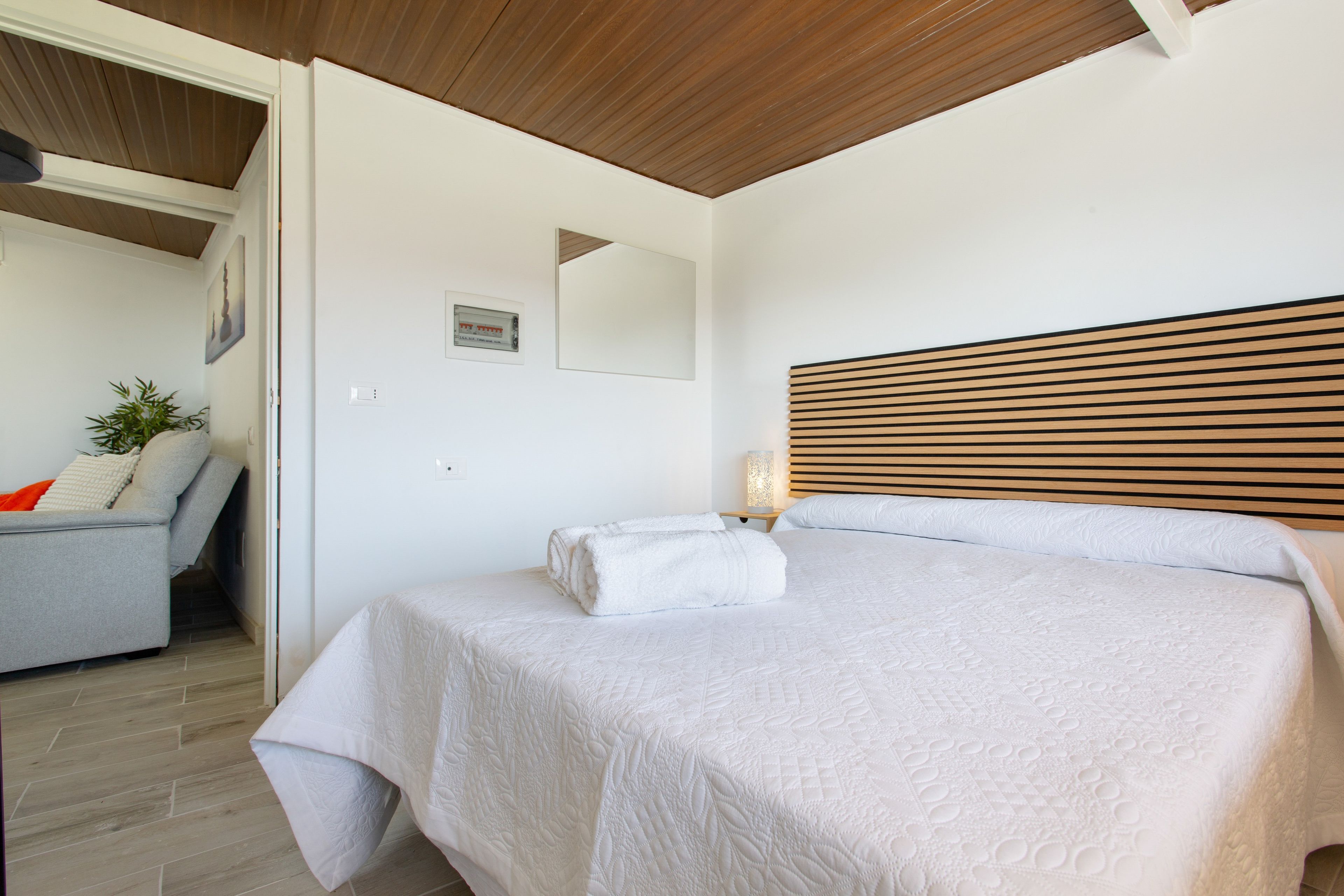 A bedroom with a bed, a nightstand, a lamp, a chair, and a wall-mounted picture.