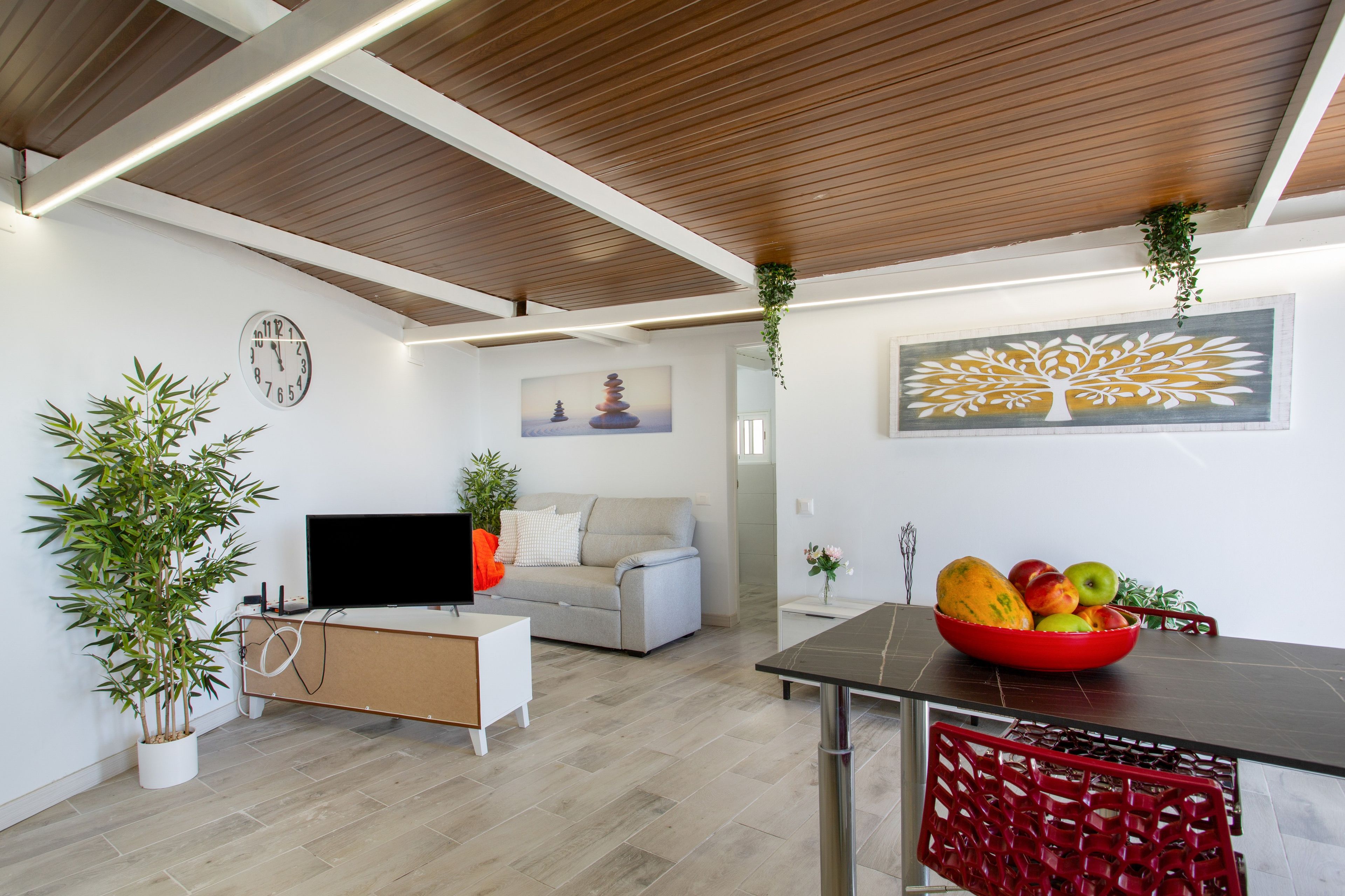 A modern living room with a flat-screen TV, a sofa, a dining table with a red basket, and a wall clock.