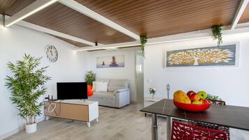 A modern living room with a flat-screen TV, a sofa, a dining table with a red basket, and a wall clock.