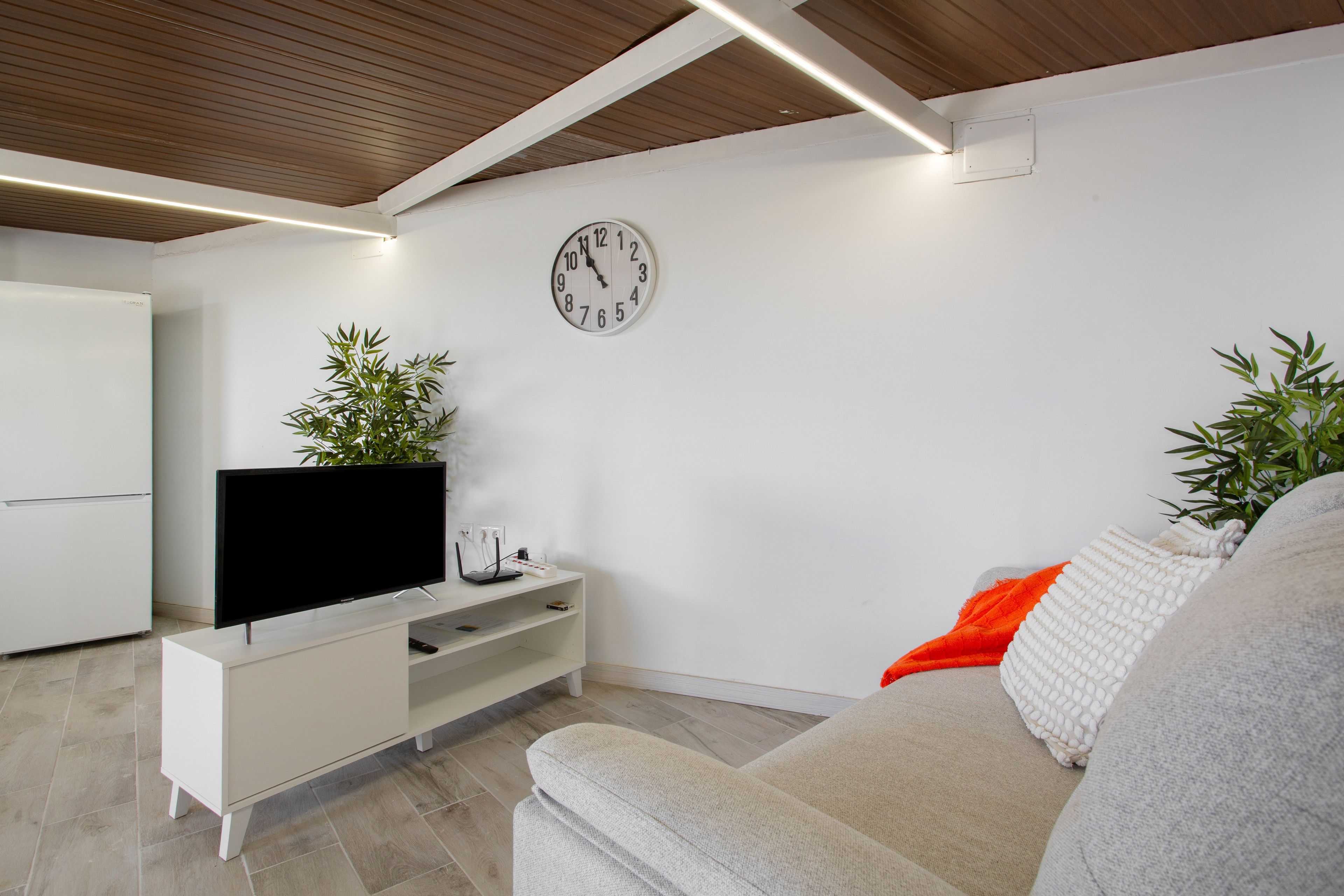 Ein modernes Wohnzimmer mit einem Flachbildfernseher auf einer weißen Unterhaltungseinheit, einer Uhr an der Wand und einem Sofa mit einer Tagesdecke.