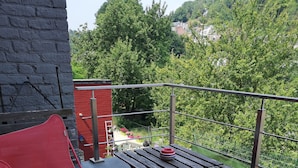 A balcony with a table and two red chairs, overlooking a lush green area with trees and a building in the background.