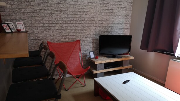 A room with a brick wall, a television on a wooden stand, a red folding chair, and a white table.