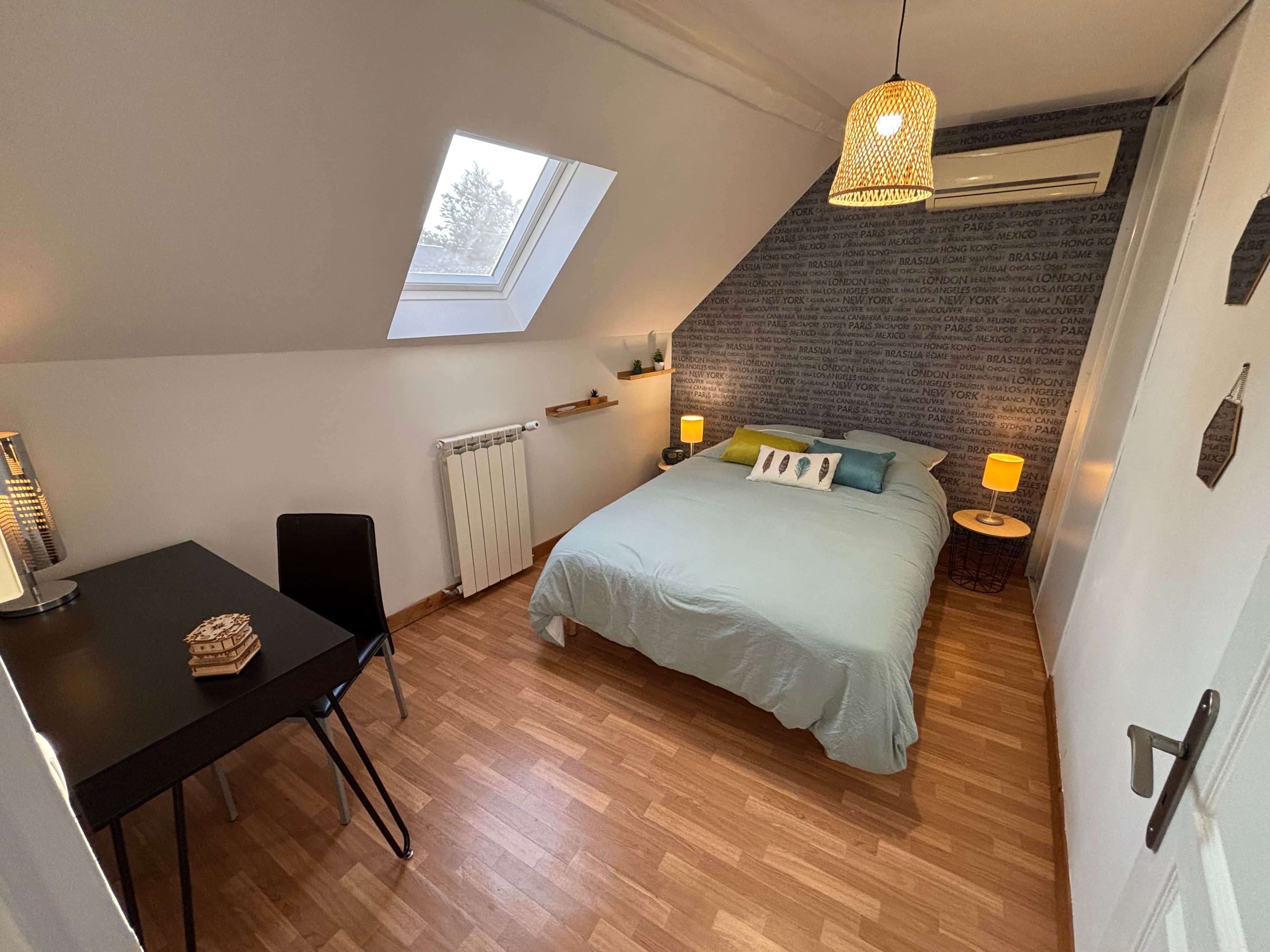A bedroom with a sloped ceiling, a skylight, a bed with pillows, a bedside table with a lamp, a radiator, and a small table with a decorative item.