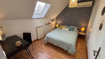 Quarto com teto inclinado, claraboia, cama com almofadas, mesa de cabeceira com abajur, radiador e uma pequena mesa com um objeto decorativo.