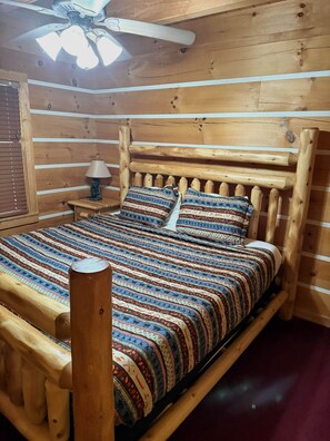 A wooden bed with a patterned bedspread and pillows, a nightstand with a lamp, and a ceiling fan. - Sugar Bear Smoky Mountain Cabin Getaway (Sevier County)