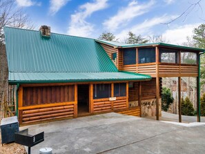 A log cabin with a green metal roof, a stone foundation, and a concrete patio.