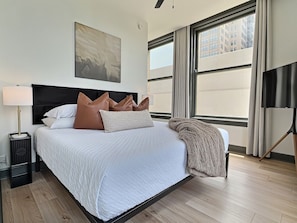 A hotel room with a large bed, a nightstand, a lamp, and a view of buildings through the window.