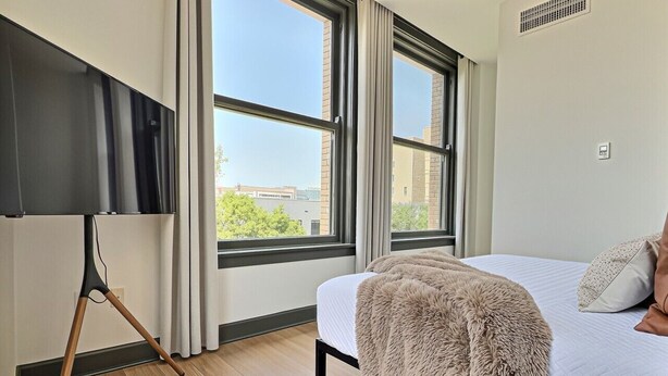 Een slaapkamer met een bed, een groot raam, een staande lamp en een uitzicht op gebouwen.