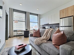 A modern living room with a grey sofa, a coffee table, and a book. There is a kitchen area with cabinets and a refrigerator. - Downtown loft near theatres and the ballpark (Birmingham)