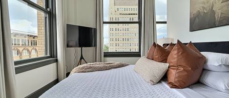 Habitación de hotel con una cama grande, una televisión de pantalla plana y vistas a un paisaje urbano desde la ventana.
